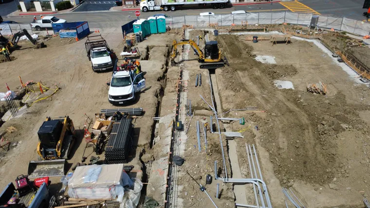 Aerial view of a deep utility excavation with conduit staged alongside an occupied commercial site